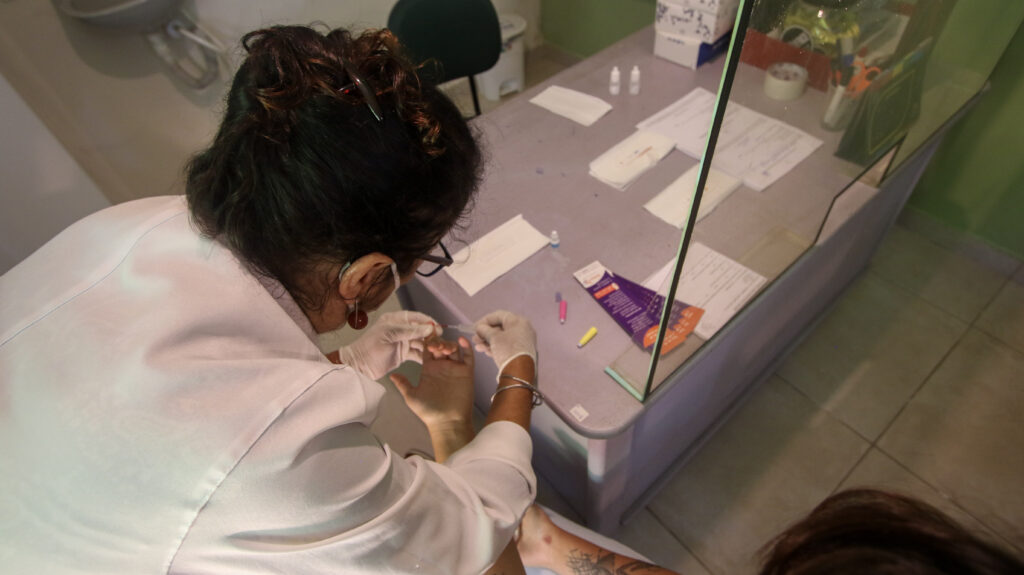 A imagem mostra uma profissional de saúde, vestindo um jaleco branco e luvas, realizando um teste rápido em uma pessoa. A cena ocorre em um ambiente clínico, com uma mesa contendo materiais médicos, como testes rápidos, álcool e documentos. A profissional está segurando a mão do paciente enquanto realiza o procedimento.